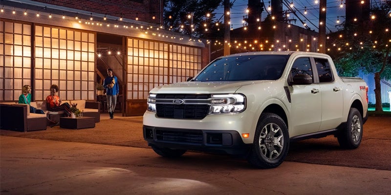 A white 2024 Ford Maverick pickup truck parked outside of a building. under a canopy of warm string lights. Friends are gathered near the truck socializing.