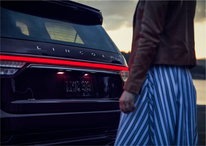 A person is shown near the rear of a 2023 Lincoln Aviator® SUV as the Lincoln Embrace illuminates the rear lights | Don Franklin Lincoln in London KY